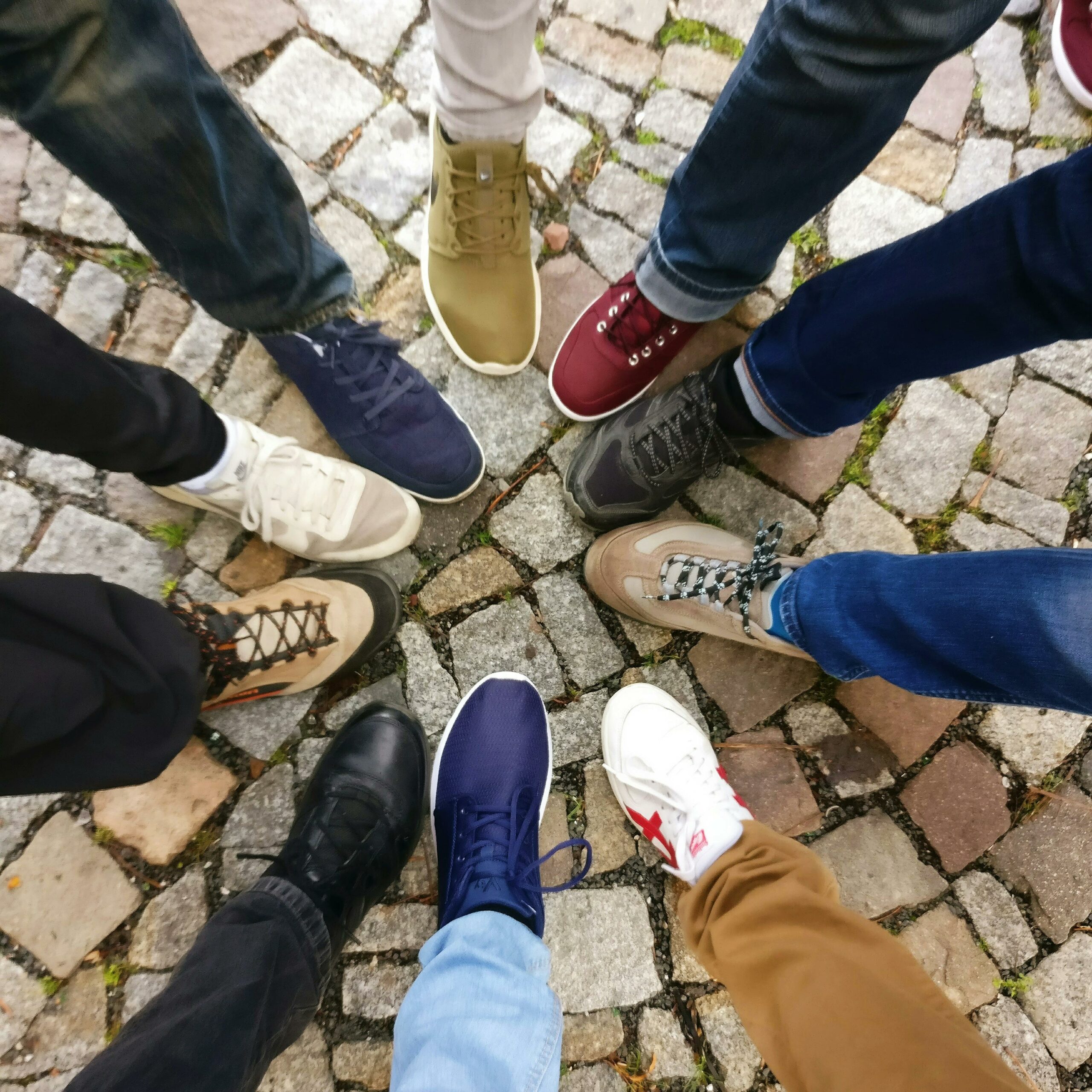 A diverse group of people's feet and legs standing in a circle, each wearing different types of shoes or sneakers.