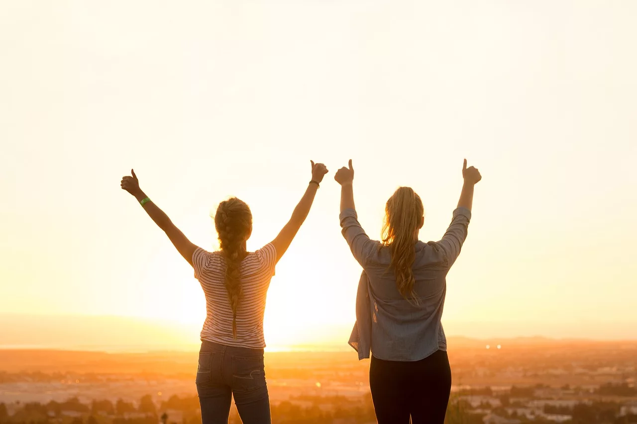 Two women standing on a beach at sunset, facing the water with their arms raised in celebration, embodying joy and triumph as they embrace the moment.